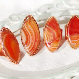 Puede incluir: Cuatro anillos de cobre con piedras de ágata de forma ovalada. Las piedras son una mezcla de naranja, rojo y blanco con un patrón de remolino.