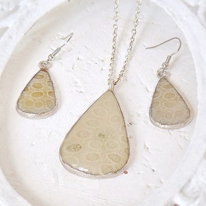 May include: A set of silver tone jewelry featuring a teardrop shaped pendant and matching earrings. The pendant and earrings are made of a light beige material with a textured pattern.