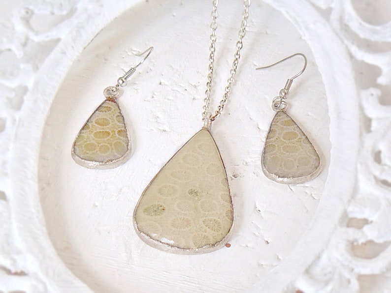 May include: A set of silver tone jewelry featuring a teardrop shaped pendant and matching earrings. The pendant and earrings are made of a light beige material with a textured pattern.