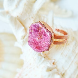 May include: A pink gemstone ring with a copper band. The ring is set on a white seashell.