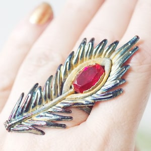 May include: A ring shaped like a feather with a red gemstone in the centre. The feather is made of metal with a gold and blue iridescent finish.