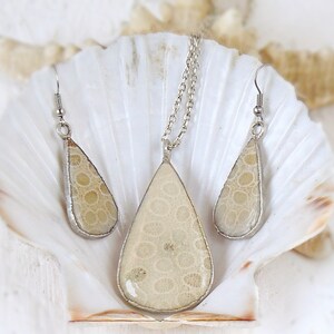 May include: A set of silver teardrop-shaped earrings and a necklace with a beige fossilized coral pendant. The jewelry is displayed on a white seashell.