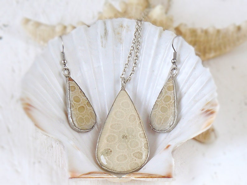 May include: A set of silver teardrop-shaped earrings and a necklace with a beige fossilized coral pendant. The jewelry is displayed on a white seashell.
