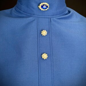 Op de afbeelding: Een close-up van een koningsblauw overhemd met een hoge kraag. De kraag heeft een blauwe ovale broche omringd door kleine heldere stenen. Het overhemd heeft drie decoratieve bloemvormige knopen aan de voorkant.
