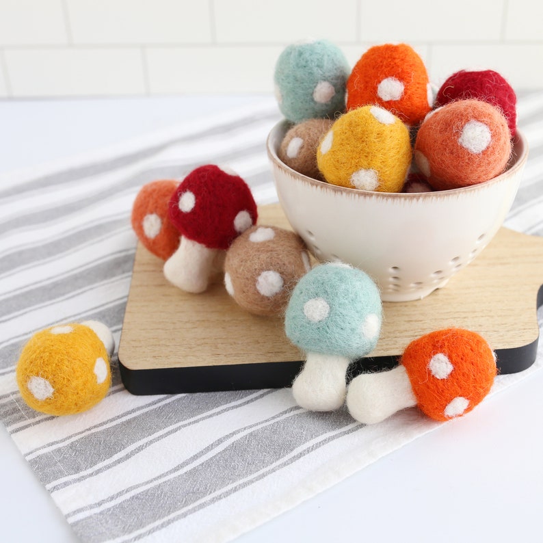 May include: A collection of felt mushroom ornaments in various colors, including red, orange, yellow, blue, and brown. The mushrooms have white polka dots and are arranged in a white bowl and on a wooden surface.