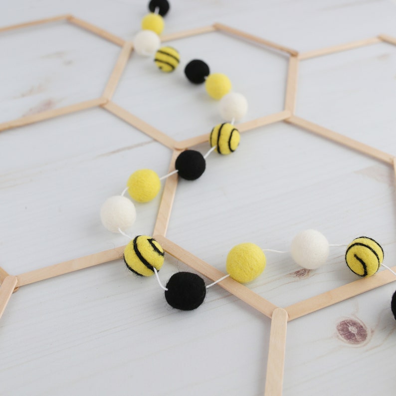 May include: A yellow and black felt bee garland hangs on a wooden honeycomb frame. The garland is made of felt balls with black stripes and white felt balls.