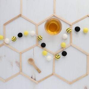 May include: A wooden honeycomb frame with a string of yellow and black felt bee ornaments and a small glass jar of honey.