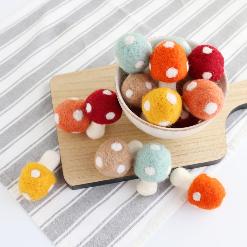 May include: A collection of felt mushroom ornaments in various colors, including red, orange, yellow, and blue. The mushrooms have white polka dots and are arranged in a bowl and on a wooden surface.