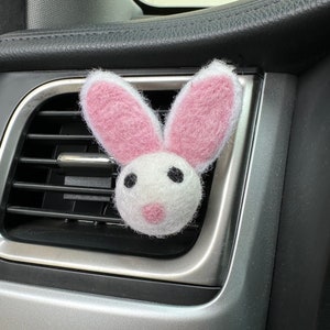 Puede incluir: Un ambientador de coche de fieltro rosa y blanco en forma de conejo que cuelga de la ventilación de un coche.