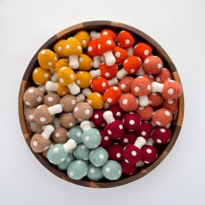 May include: A bowl filled with felt mushroom ornaments in various colors, including red, orange, yellow, and green. The mushrooms have white polka dots and are arranged in a circular pattern.