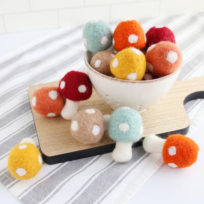 May include: A collection of felt mushrooms in various colors, including red, orange, yellow, and green, with white polka dots. The mushrooms are arranged in a white bowl and on a wooden surface.