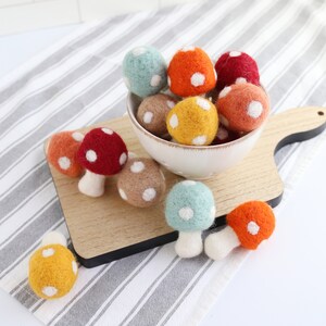 May include: A bowl filled with felt mushroom ornaments in various colors, including red, orange, yellow, green, and brown. The mushrooms have white polka dots and are arranged on a wooden cutting board.