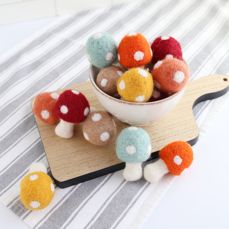 May include: A bowl filled with felt mushroom ornaments in various colors, including red, orange, yellow, green, and brown. The mushrooms have white polka dots and are arranged on a wooden cutting board.