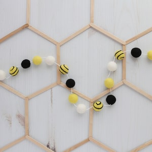 May include: A garland of yellow and black felt balls with a bee pattern, strung on white thread. The garland is hanging on a wooden honeycomb background.