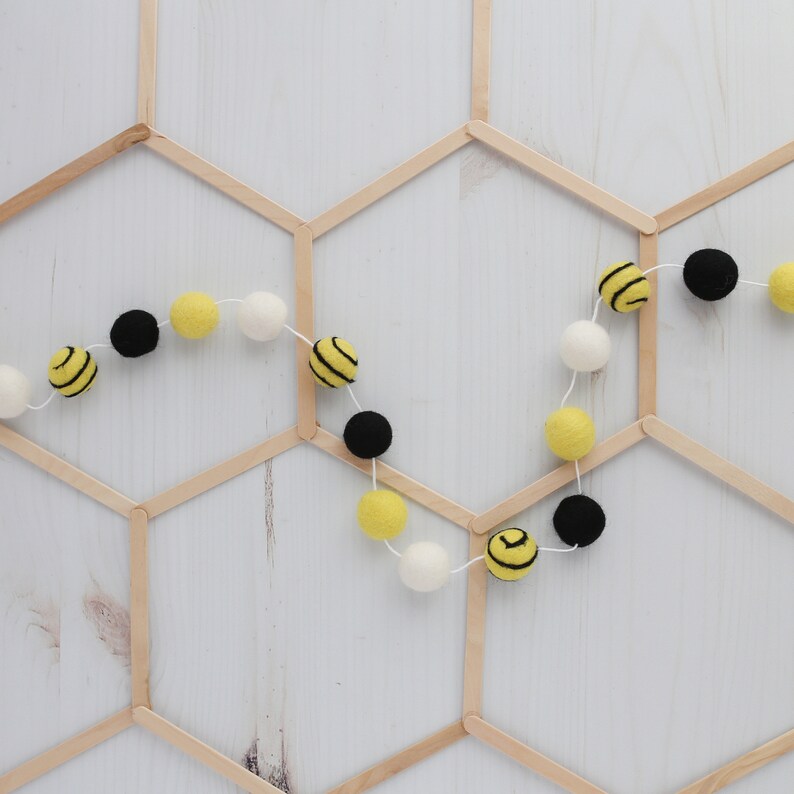 May include: A garland of yellow and black felt balls with a bee pattern, strung on white thread. The garland is hanging on a wooden honeycomb background.