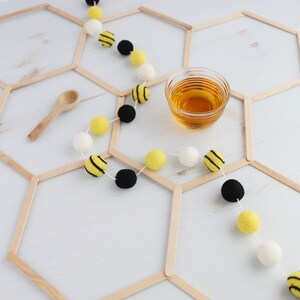 May include: A white background with a honeycomb pattern made from wooden craft sticks. A string of yellow and black pom-poms with white pom-poms in between is draped over the honeycomb pattern. A small wooden spoon and a glass cup of honey are also on the background.