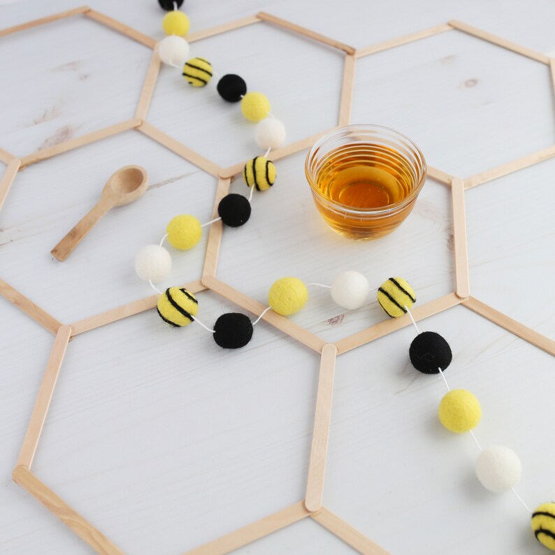 May include: A white background with a honeycomb pattern made from wooden craft sticks. A string of yellow and black pom-poms with white pom-poms in between is draped over the honeycomb pattern. A small wooden spoon and a glass cup of honey are also on the background.