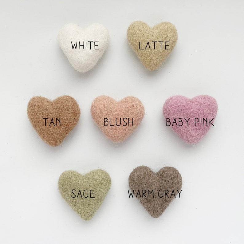 May include: A collection of heart-shaped felt ornaments in various colors, including white, latte, tan, blush, baby pink, sage, and warm gray. Each heart has a color name printed on it.