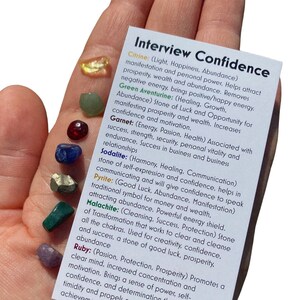 May include: A close-up of a hand holding a collection of crystals with a card that reads "Interview Confidence" and lists the names and properties of each crystal. The crystals are citrine, green aventurine, garnet, sodalite, pyrite, malachite, and ruby.