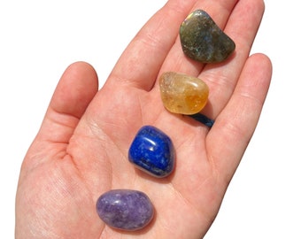 May include: Four tumbled stones in a hand. The stones are a dark green labradorite, a yellow citrine, a blue lapis lazuli, and a purple amethyst.