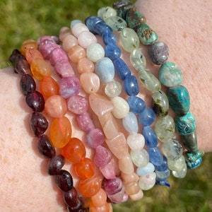 Op de afbeelding: Een verzameling edelsteenarmbanden in verschillende kleuren, waaronder tinten rood, oranje, roze, blauw en groen. De armbanden zijn gemaakt van gepolijste, onregelmatig gevormde stenen die aan elkaar zijn geregen. De armbanden worden om een pols getoond.
