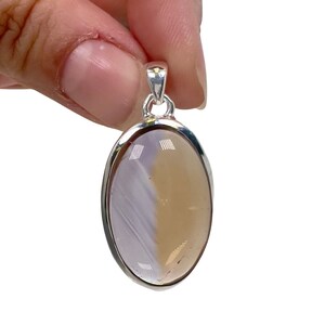 May include: Oval pendant with a silver-colored bail. The pendant features a two-toned stone, with a light purple and tan color scheme. The stone is set in a silver-colored metal frame. The pendant is held by a hand.