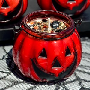 May include: Three red glass candle holders shaped like jack-o'-lanterns with carved triangular eyes and wide smiling mouths. Each container features a metal handle and is filled with wax topped with various dark botanical elements and a wick.
