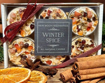 May include: A box set of six tea light candles with the text "Winter Spice" and "All Natural Candle". The candles are decorated with colorful stones and spices. Cinnamon sticks and dried orange slices are also visible.