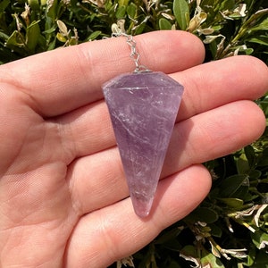 May include: A purple amethyst crystal pendulum with a silver chain. The pendulum is in the shape of a point and is held in a hand.