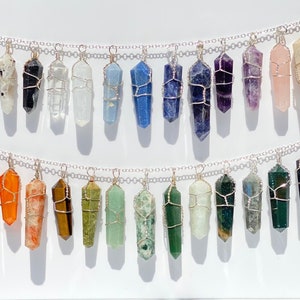 Könnte beinhalten: Eine Sammlung von 24 drahtgewickelten Kristallspitzenanhängern in verschiedenen Farben, darunter klar, schwarz, blau, lila, rosa, orange, braun, grün und grau. Die Anhänger sind in zwei Reihen auf einem weißen Hintergrund angeordnet.