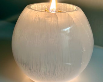 Può includere: Un portacandele in selenite bianco con una candela accesa all'interno. Il portacandele è rotondo e ha una superficie liscia e lucida.