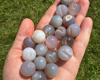 May include: A collection of 18 gray agate spheres with white and gray banding. The spheres are smooth and polished, and they are arranged in a pile on a hand.