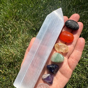 May include: A selenite wand with a pointed tip, lying on a hand with seven tumbled chakra stones. The stones are black, orange, yellow, green, blue, purple, and clear.