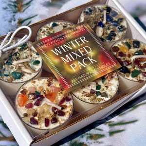 Puede incluir: Un conjunto de seis velas redondas en una caja blanca, cada una cubierta con varios cristales coloridos y hierbas secas. Una etiqueta cuadrada en el centro dice New Moon Beginnings Winter Mixed Pack All Natural Candles. Un cordel blanco rodea la caja.