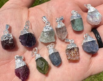 Può includere: Una collezione di 11 ciondoli in cristallo grezzo, ognuno con una ghiera in argento. I cristalli sono di vari colori, tra cui verde, blu, viola, rosa e trasparente.