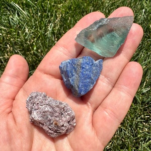 May include: Three rough, natural gemstones: a green fluorite, a blue lapis lazuli, and a pink lepidolite.