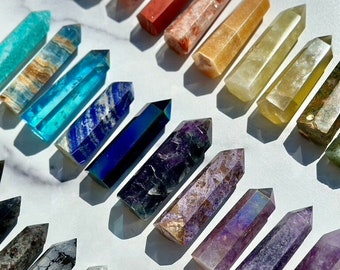 May include: A collection of polished crystal wands in various colors, including turquoise, blue, purple, and pink. Each wand has a pointed tip and a rectangular base. The crystals are arranged on a white surface, with shadows adding depth.