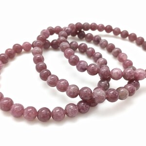 Puede incluir: Tres pulseras apiladas hechas de cuentas de lepidolita rosa. Las cuentas son redondas y tienen una superficie ligeramente texturizada.