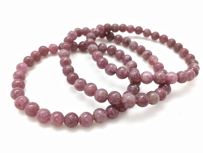 Puede incluir: Tres pulseras apiladas hechas de cuentas de lepidolita rosa. Las cuentas son redondas y tienen una superficie ligeramente texturizada.