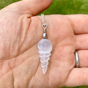 K&ouml;nnte beinhalten: Ein wei&szlig;er Selenit-Kristall-Anh&auml;nger in Form einer Spirale, der an einer silbernen Kette h&auml;ngt. Der Anh&auml;nger wird in einer Hand gehalten.
