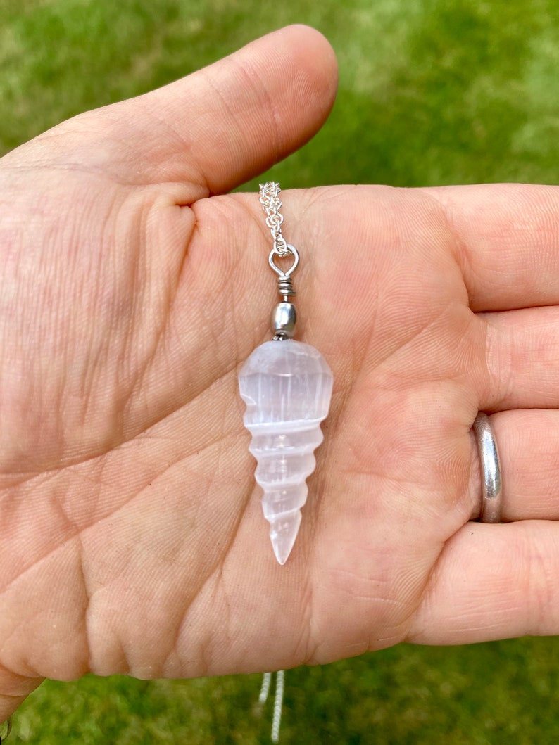 K&ouml;nnte beinhalten: Ein wei&szlig;er Selenit-Kristall-Anh&auml;nger in Form einer Spirale, der an einer silbernen Kette h&auml;ngt. Der Anh&auml;nger wird in einer Hand gehalten.
