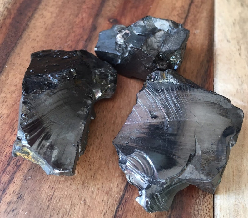 Pu&ograve; includere: Tre pezzi di vetro vulcanico grezzo di ossidiana. Le pietre scure e riflettenti hanno bordi frastagliati e un aspetto nero lucido. Sono esposte su una superficie di legno.