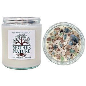 May include: A white candle with a clear glass jar and a label that reads "New Moon Beginnings Mother Nature All Natural Candles". The candle is decorated with small pieces of green, brown, and white stones.