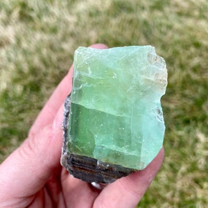 May include: A large, green, translucent crystal with a rough, natural texture. The crystal has a layered appearance with darker green and brown bands.