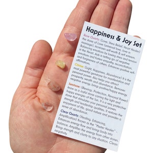 May include: A set of five small crystals, including rose quartz, citrine, sunstone, and clear quartz. The crystals are arranged on a hand, with a card describing their properties and uses. The card reads "Happiness & Joy Set".