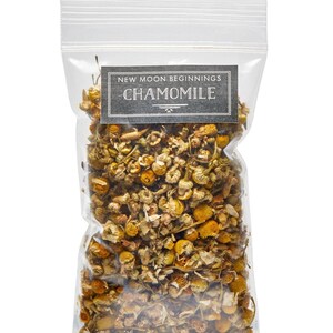 May include: A clear plastic bag filled with dried chamomile flowers. The bag has a rectangular label with the text "NEW MOON BEGINNINGS CHAMOMILE" in a dark gray font. The flowers are a mix of yellow and brown.