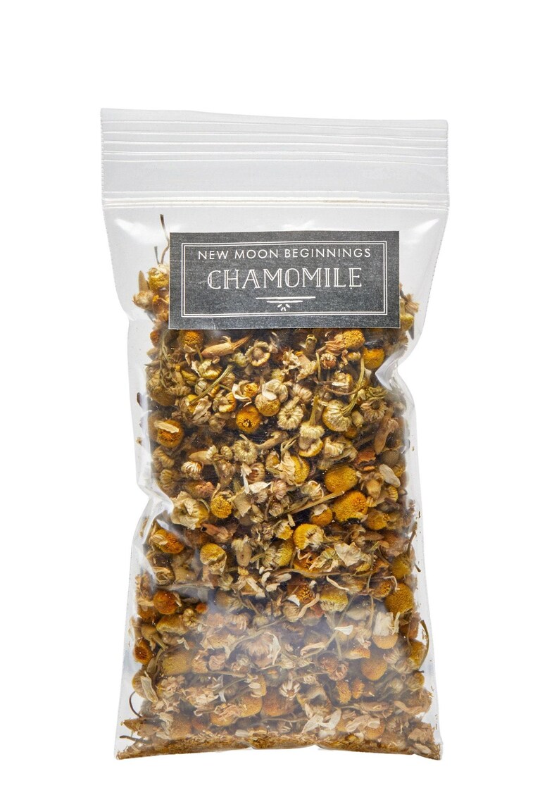 May include: A clear plastic bag filled with dried chamomile flowers. The bag has a rectangular label with the text "NEW MOON BEGINNINGS CHAMOMILE" in a dark gray font. The flowers are a mix of yellow and brown.