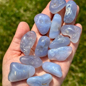 May include: A handful of polished, smooth, blue chalcedony stones. The stones vary in size and shape, with a translucent quality and subtle banding. The background is blurred green grass, highlighting the stones' natural beauty.