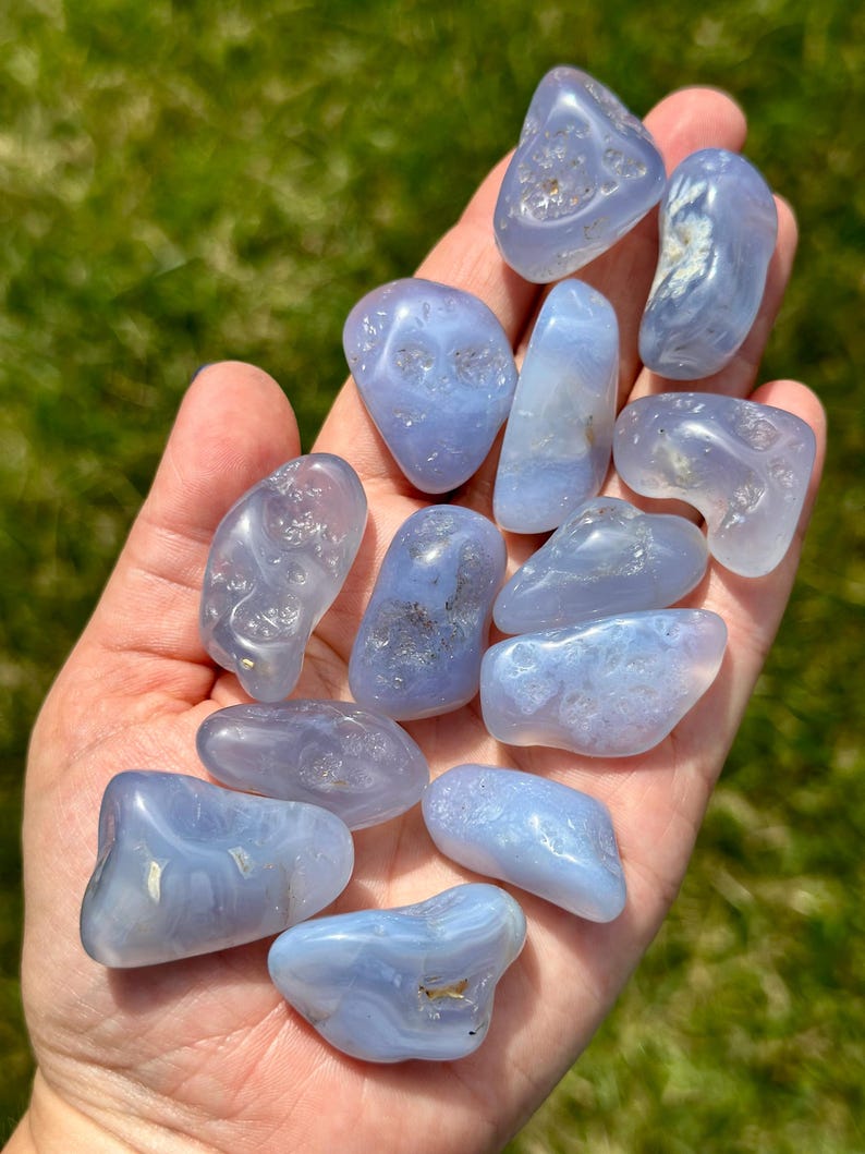 May include: A handful of polished, smooth, blue chalcedony stones. The stones vary in size and shape, with a translucent quality and subtle banding. The background is blurred green grass, highlighting the stones' natural beauty.