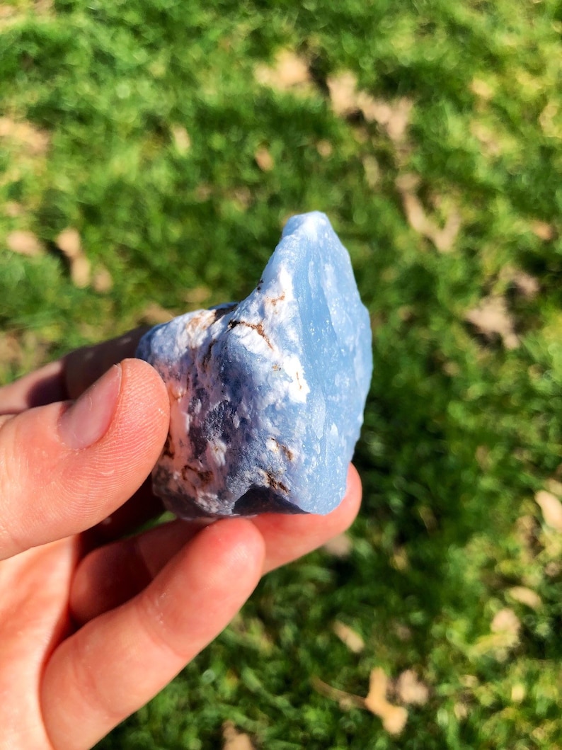 May include: A light blue, rough-textured, natural stone with white and brown markings. The stone is being held in a hand.
